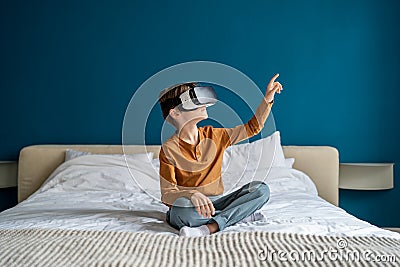 Child boy wearing virtual reality headset sitting on bed at home exploring metaverse. VR concept Stock Photo