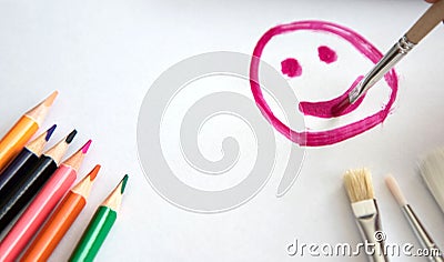 A child with a brush and raspberry paint draws a happy smile or smiling face on a sheet of white paper, on the sheet are colored Stock Photo