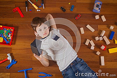 Child with 3D Virtual Reality, VR cardboard glasses lying on wooden floor. Many toys around him Stock Photo