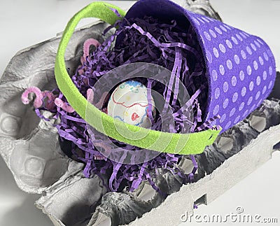 A polkadot purple Easter basket with lime green handle rests on an open empty egg carton. See one egg and paper grass. Stock Photo