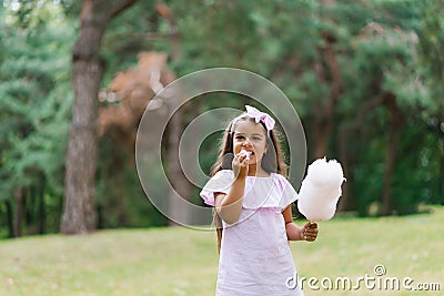 Child girl eats cotton candy on a sunny summer day and licks sticky fingers. Walk with children in the park on vacation Stock Photo