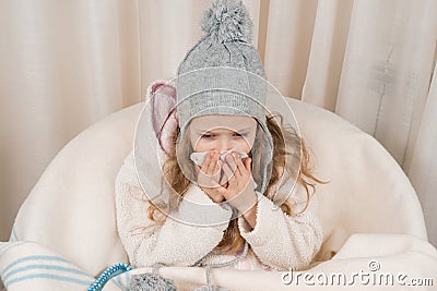 Child girl at home in chair with warm knitted hat woolen blanket sneezes in handkerchief. Season autumn winter colds Stock Photo