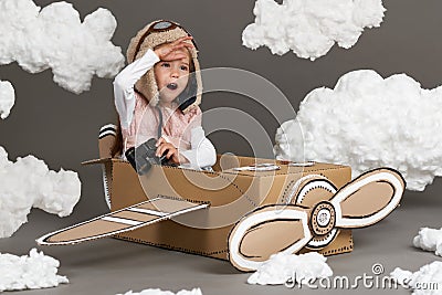 The child girl plays in an airplane made of cardboard box and dreams of becoming a pilot, clouds of cotton wool on a gray backgrou Stock Photo