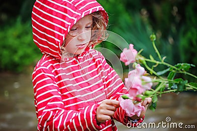 Child girl in red striped raincoat playing with wet roses in rainy summer garden. Nature care concept. Stock Photo