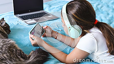 A child with headphones playing a computer game on the phone. A little girl lies on the bed with a laptop and a Stock Photo