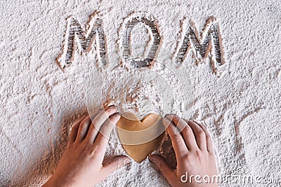 Child holding heart shaped cookie and word mom written in flour, Mothers day concept Stock Photo