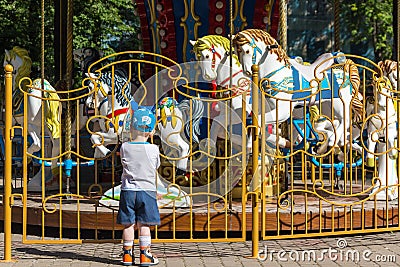 The child looks at old French carousel in a holiday park. Horses on a traditional fairground vintage carousel. Stock Photo