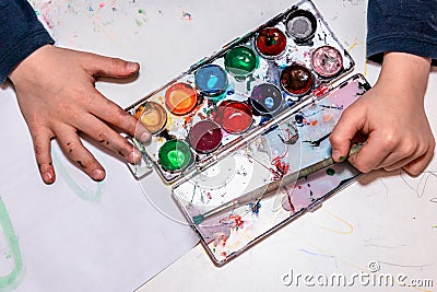Child messy hands paint with watercolors, palette with acrylic paints for drawing on the white table, bright colorful abstract Stock Photo