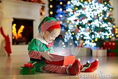 Child opening present at Christmas tree at home. Kid in elf costume with Xmas gifts and toys. Little baby boy with gift box and Stock Photo