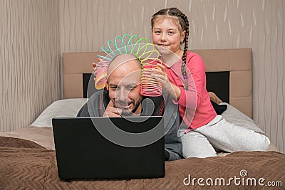 A child prevents a man from working on a laptop in his bedroom. The daughter puts the slinky toy to the father`s head. Family is Editorial Stock Photo