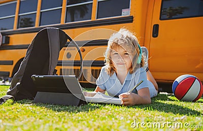 Child pupil does school homework laying on grass in the park near school bus. School kid outdoor. Stock Photo