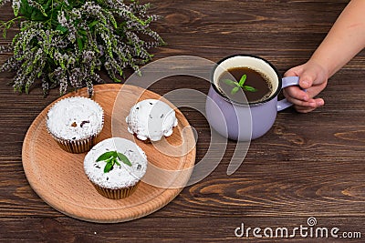 A child`s hand holds a cup of tea and mint near the board with muffins with black currant, on a background of flowers Stock Photo