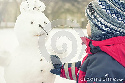 A child sculpts a snowman in the winter on the street, a boy in a red overall Stock Photo