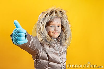 The child shows a thumb up. Happy cute little girl in a down jacket on a yellow background. Winter concept. Stock Photo