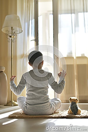 The child sits with his back at home. On the mat with a plush toy dog. Meditation in a yoga pose with hands makes mudra Stock Photo