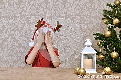 A child in a striped cap with deer horns, hid behind his hands and smiles, waiting for a surprise Stock Photo