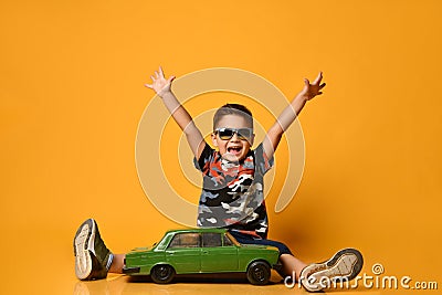 Child in sunglasses, camouflage t-shirt. Raised hands, sitting on floor near green model of retro car, posing on orange background Stock Photo