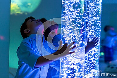 Child in therapy sensory stimulating room, snoezelen. Child interacting with colored lights bubble tube lamp during therapy. Stock Photo