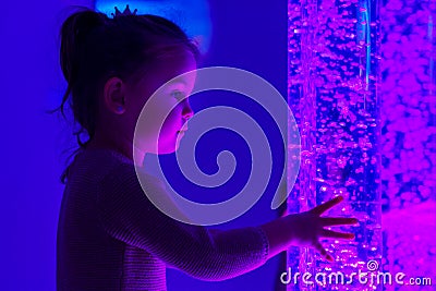 Child in therapy sensory stimulating room, snoezelen. Child interacting with colored lights bubble tube lamp during therapy. Stock Photo