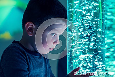 Child in therapy sensory stimulating room, snoezelen. Child interacting with colored lights bubble tube lamp during therapy. Stock Photo