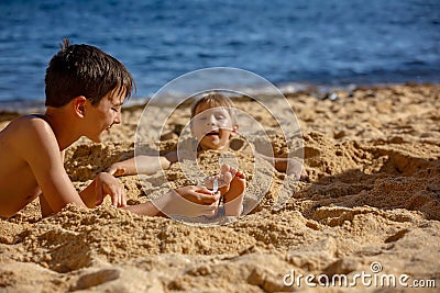 Child, tickling sibling on the beach on the feet with feather, kid cover in sand, smiling, laughing Stock Photo