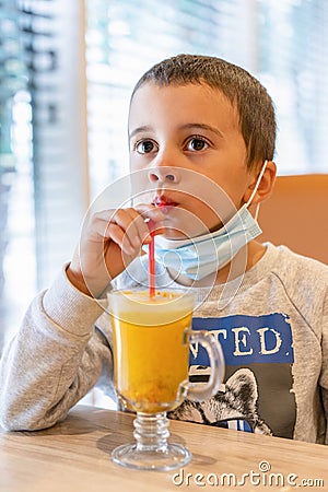 A child wearing a mask in a cafe during a pandemic. A boy in a cafe drinks sea buckthorn tea in a cafe during a pandemic Stock Photo