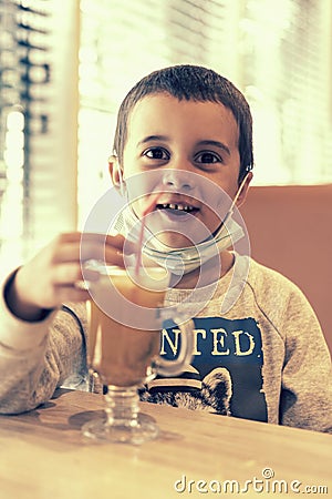 A child wearing a mask in a cafe during a pandemic. A boy in a cafe drinks sea buckthorn tea in a cafe during a pandemic. toned Stock Photo