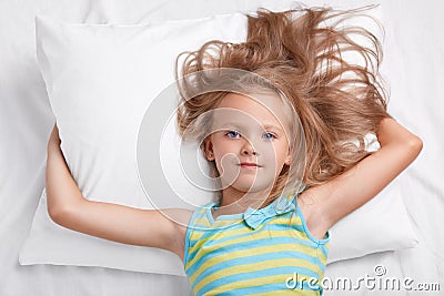Children, awakening and bed time concept. Little adorable relaxed girl, dressed in casual outfit, feels relaxed in comfortable bed Stock Photo