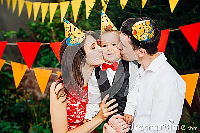 Children birthday party. Family father and mother hold son of one year of birth in arms and kiss cheekin the background of park an Stock Photo