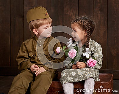 Children boy are dressed as soldier in retro military uniforms and girl in pink dress sitting on old suitcase, dark wood backgroun Stock Photo