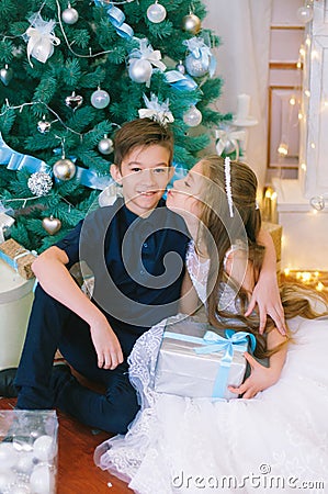 Children, a boy and a girl in a white ball gown near a Christmas tree in holiday dresses with gifts and silver confetti. Stock Photo