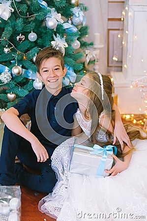 Children, a boy and a girl in a white ball gown near a Christmas tree in holiday dresses with gifts and silver confetti. Stock Photo