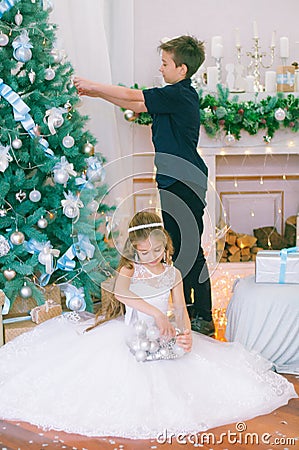 Children, a boy and a girl in a white ball gown near a Christmas tree in holiday dresses with gifts and silver confetti. Stock Photo