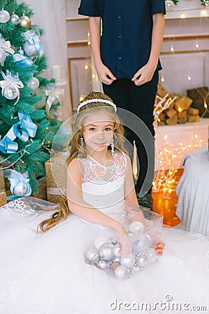 Children, a boy and a girl in a white ball gown near a Christmas tree in holiday dresses with gifts and silver confetti. Stock Photo