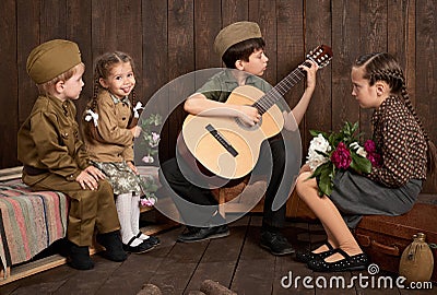 Children are dressed in retro military uniforms sitting and playing guitar, sending a soldier to the army, dark wood background, r Stock Photo