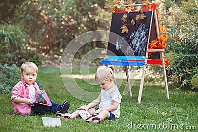 Children kids, boy and girl sitting in grass outside by drawing easel with books reading studying learning Stock Photo