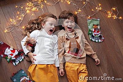 Children lying, laughing, have fun with Christmas decor, garland of light bulbs. Beautiful kids wearing sweater with deer Stock Photo