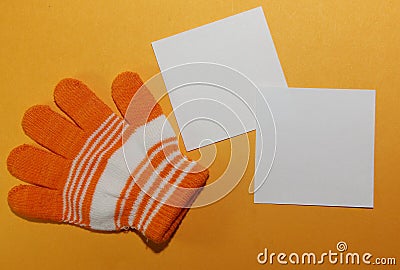 Children`s glove, on the child`s hand, orange striped lies on a yellow surface with two white square shaped blank sheets for writi Stock Photo