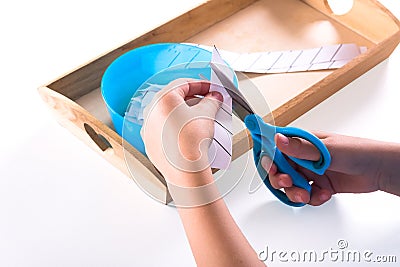 Children`s hands hold blue scissors and cut the paper. On a wooden tray are Montessori materials for a lesson Stock Photo