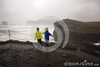 Children, standing at the edge of the ocean on heavy rain day near Dyrholaey, watching the huge waves, Iceland wintertime Stock Photo