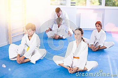 Children in white kimono sits in butterfly pose and practices stretching in sport gym Stock Photo