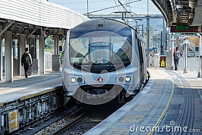 Fast metro train arrives at open-air station, people walk on platform. MTR Corporation. Editorial Stock Photo