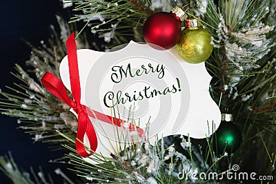 Christmas Card with Merry Christmas message tied with a red bow, in a snowy tree with red and green ornaments. It`s a closeup ph Stock Photo