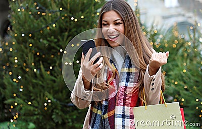 Christmas girl. Happy excited young woman with shopping bags in her hand buying christams presents with her smart phone outdoors Stock Photo