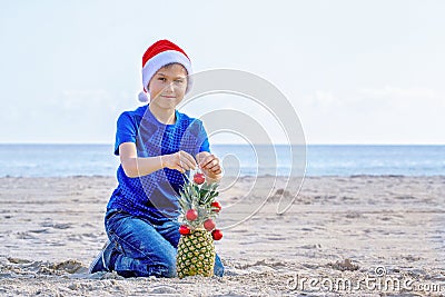 Christmas holiday. Boy in red Santa hat decorating pineapple as a Christmas tree on a sunny sandy beach by the sea Stock Photo