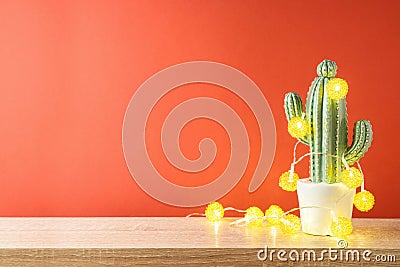 Christmas holiday greeting card with cactus as alternative Christmas tree and lights garland on wooden shelf over red background Stock Photo