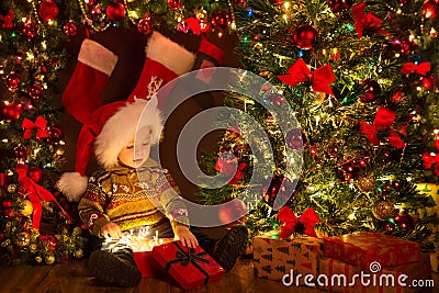 Christmas Kid opening Gift Box with Xmas Lights Garland. Cute Baby in Santa Hat sitting next to Decorated Fir Tree in Dark Room Stock Photo