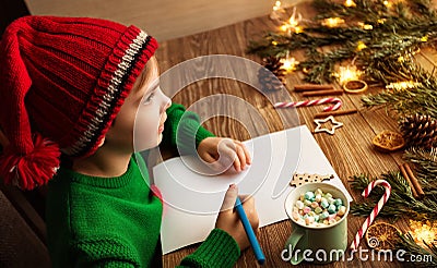 Christmas Kid writing Letter to Santa. Happy Child in Red Santa Hat dreaming about Presents at Home. Xmas Gifts Shopping Wish List Stock Photo