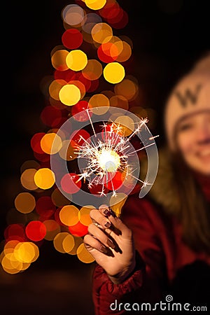 Christmas photography. the girl holds a sparkler in her hand, the lights of garlands on the tree are visible from behind. Stock Photo