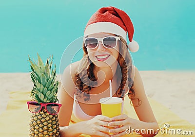 Christmas portrait of happy smiling young woman drinking fresh juice in red santa hat with pineapple lying on beach over blue Stock Photo